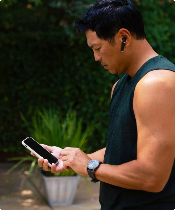 A man wearing earbuds, focused on his phone screen, with trees in the background.