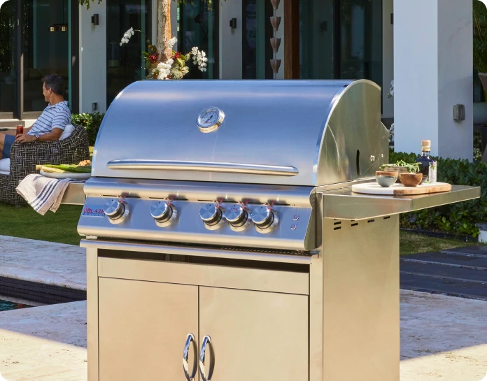 A stainless steel outdoor grill with two burners, with a relaxing outdoor zone in the background.